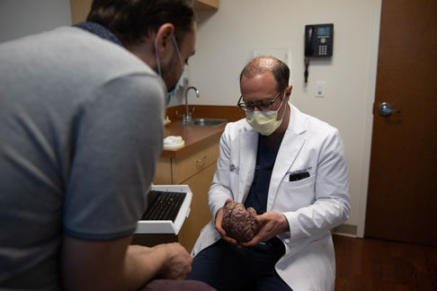 A male doctor examines a medical model of a brain with a patient in an examination room.